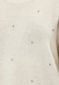 Maglione a maglia color crema con una texture morbida, caratterizzato da piccole decorazioni metalliche distribuite uniformemente sulla parte anteriore.