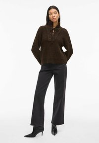 Brauner Strickpullover mit hohem Kragen und Schnürdetails auf der Vorderseite, kombiniert mit hochtaillierten schwarzen weit geschnittenen Jeans und spitzen schwarzen Schuhen.