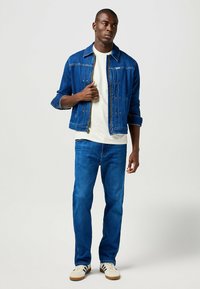 Chaqueta de mezclilla en azul oscuro con cremallera, camiseta blanca debajo, vaqueros azules y zapatillas blancas con rayas negras. Ajuste casual, detalles mínimos.