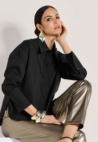 Eine Frau mit glatten, dunklen Haaren trägt große geometrische Ohrringe, ein schwarzes, strukturiertes Shirt, metallisch goldene Hosen und klobige Armbänder aus Silber und Gold.