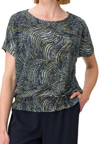 Femme portant un chemisier à manches courtes avec un motif circulaire abstrait noir, blanc et vert olive et un pantalon foncé, main dans la poche.