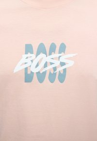 Chemise rose clair avec le mot « BOSS » écrit deux fois ; grandes lettres bleues en arrière-plan et lettres blanches stylisées en superposition.