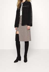 Donna che indossa un giubbotto nero con bottoni e occhielli metallici, una maglia bianca, una gonna a quadretti con spacco all'altezza della coscia e stivali neri fino al ginocchio.
