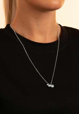 Silberne Kettenhalskette mit drei klaren Edelsteinen, die über einem schwarzen T-Shirt liegt. Einfaches Design mit glatter Textur und leichtem Glanz.