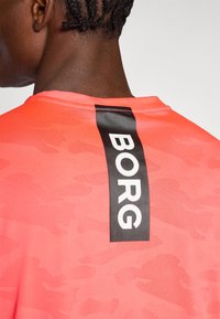 Camisa deportiva con patrón de camuflaje rojo y una etiqueta vertical negra que dice "BORG" en la parte posterior. La textura parece suave y transpirable.