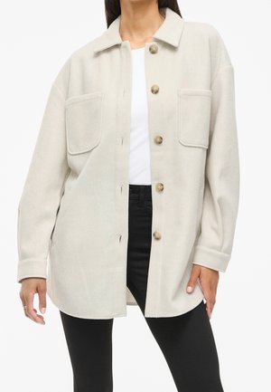 Femme portant une surchemise beige surdimensionnée à boutons avec deux poches avant, associée à un haut blanc et un pantalon noir.