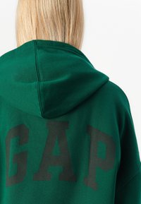 Sweat à capuche vert avec un grand logo noir "GAP" au dos, fabriqué en coton doux, avec un cordon de serrage à la capuche et des poignets côtelés.