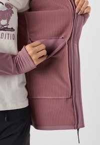 Person trägt ein helles Hemd und hält eine offene, mauvefarbene Fleecejacke mit Rippstruktur, die eine große Innentasche mit Klappe auf der linken Seite zeigt.