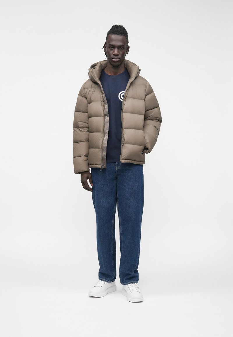 Mann steht vor weißem Hintergrund, trägt eine beige gesteppte Jacke, einen marineblauen Pullover mit rundem Logo, blaue Jeans und weiße Sneakers.