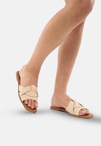 Sandales plates beiges avec des lanières croisées, fabriquées en matériau synthétique lisse. Conçues avec une semelle plate et un bout ouvert pour permettre une meilleure circulation de l'air.