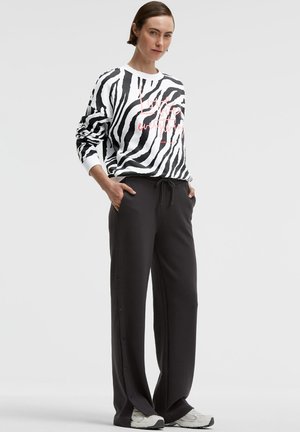 Frau trägt schwarze Hosen mit weitem Bein und seitlichen Knöpfen, weiße Sneaker und ein schwarz-weißes Sweatshirt mit Zebramuster und pinkem Text.