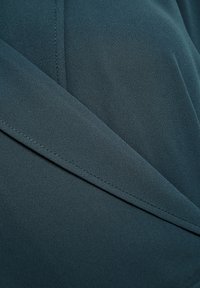 Donkerblauwgroene stof met een gladde textuur, gekenmerkt door diagonale plooien en stiksels. Het ontwerp creëert een overlappend gelaagd effect.