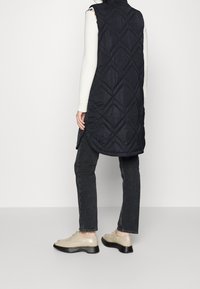 Schwarze wattierte Weste mit diamantförmigem Steppmuster, hohem Kragen und asymmetrischem Saum, getragen über einem weißen Pullover und kombiniert mit dunklen Jeans und hellen Schuhen.