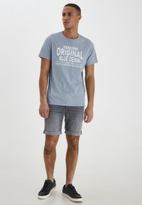 Lys blå bomull T-skjorte med hvit tekst, sammen med grå denimshorts og svarte joggesko, som viser et avslappet antrekk.