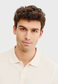 Beżowa koszulka polo o miękkiej teksturze, z złożonym kołnierzykiem i trzema guzikami, noszona przez młodego mężczyznę z kręconymi ciemnymi włosami, na neutralnym tle.