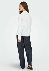 Chemise blanche à manches longues avec détails de boutons au col, associée à un pantalon large bleu foncé. Tissu lisse, coupe décontractée et design minimaliste.