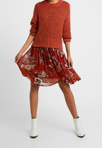 Personne portant un pull en maille couleur rouille, une jupe florale rouge et des bottines blanches, tenant la jupe sur les côtés.