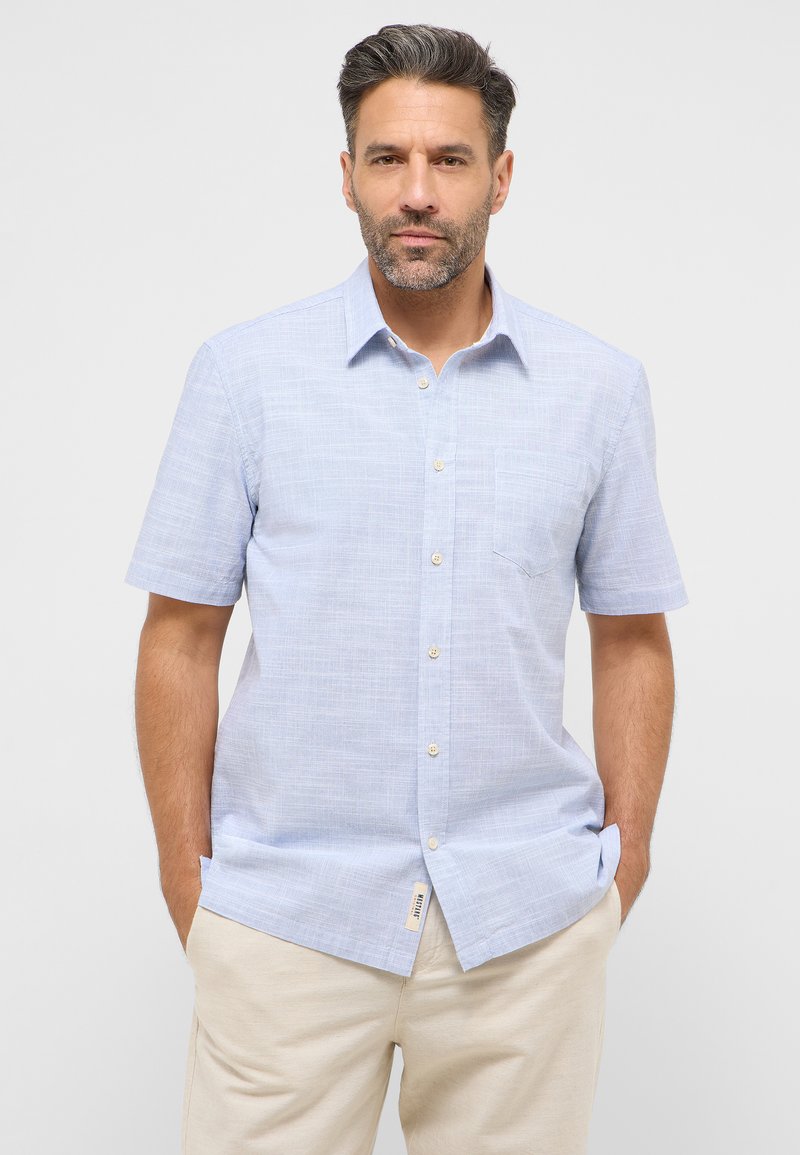 Jasnoniebieska koszula z krótkim rękawem, zapinana na guziki, z delikatnym wzorem teksturowym, pojedynczą kieszenią na piersi oraz prostym dołem. W zestawie z beżowymi spodniami.