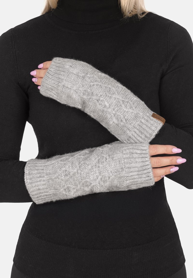 Guantes sin dedos grises de punto con patrón de trenzas, usados con los brazos cruzados sobre un suéter de cuello alto negro.