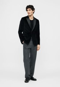 Junger Mann trägt schwarzen Samtsakko, schwarzes Hemd, graue Anzughose und schwarze elegante Schuhe, steht mit den Händen in den Taschen.