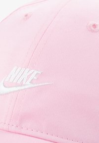 Boné rosa feito de algodão com uma superfície texturizada. Apresenta o logótipo "Nike" bordado em branco e um buraco de ventilação costurado na parte superior.
