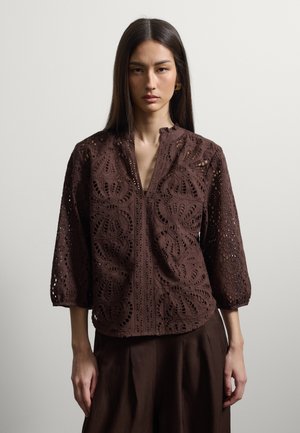 Femme aux cheveux lisses et foncés porte un chemisier en dentelle marron avec des motifs floraux découpés et une jupe assortie, debout sur un fond clair uni.