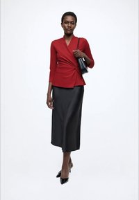 Rotes Wickel-Oberteil mit V-Ausschnitt und dreiviertel Ärmeln, kombiniert mit einem schwarzen, satinähnlichen knielangen Rock. Modell hält eine schwarze Handtasche.