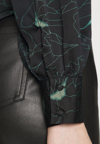 Svart blus med grönt blommönster; har samlade ärmar med knappdetalj vid manschetterna, ihop med ett svart läderplagg.