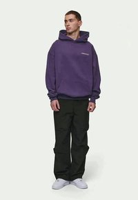 Felpa di cotone viola con tasca frontale e logo, abbinata a pantaloni cargo neri e sneakers bianche. Modello in piedi davanti a uno sfondo neutro.