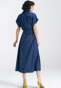 Jeansowa sukienka midi z krótkimi rękawami w stylu dolman, zebranym w talii i plisami z tyłu. Połączona z jasnoniebieskimi sandałami na wysokim obcasie.