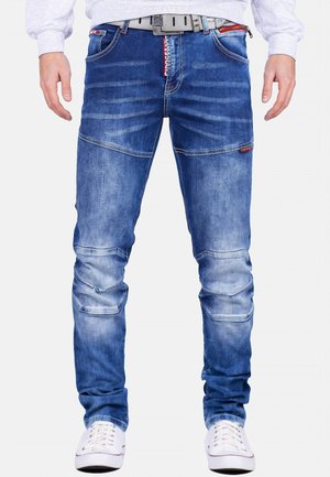 Mand iført blå falmede jeans med et gråt bælte og hvide sneakers, stående med armene afslappet ned langs siderne.