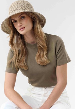 Woman with long blonde hair wearing a beige woven hat, olive green short-sleeve shirt, and white pants, posed against a plain light background.