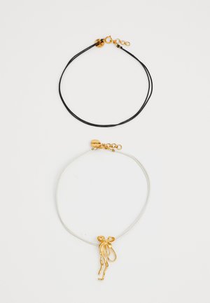 Dos collares: un cordón negro con un cierre dorado y un cordón blanco que presenta un colgante abstracto dorado que se asemeja a cintas ondulantes.