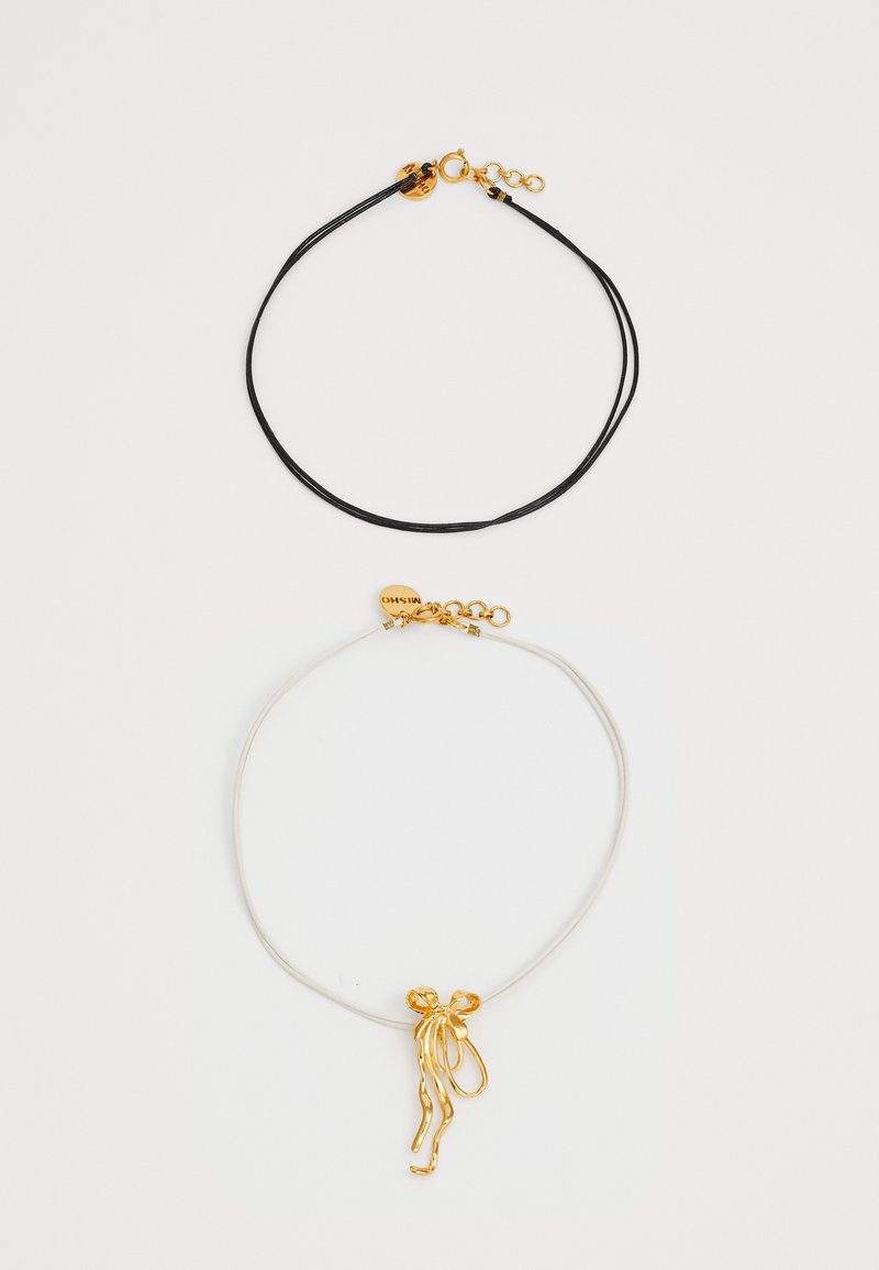 Due collane: una corda nera con un fermaglio dorato e una corda bianca con un pendente astratto dorato che assomiglia a nastri fluttuanti.