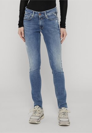 Jean bleu slim avec une décoloration subtile, porté avec des baskets blanches et un top noir à manches longues, debout devant un fond clair uni.