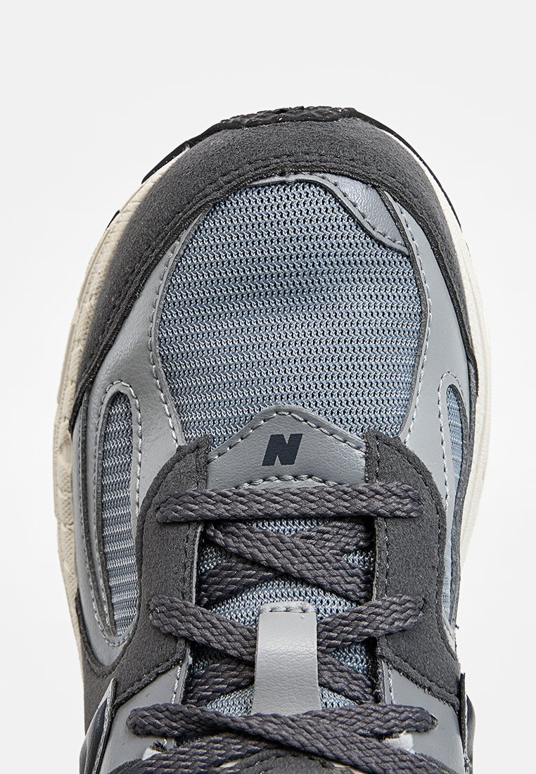 Zapatilla de malla gris y ante con una superficie texturizada, que cuenta con una lengüeta acolchada, cordones grises y un logo visible cerca del dedo del pie.