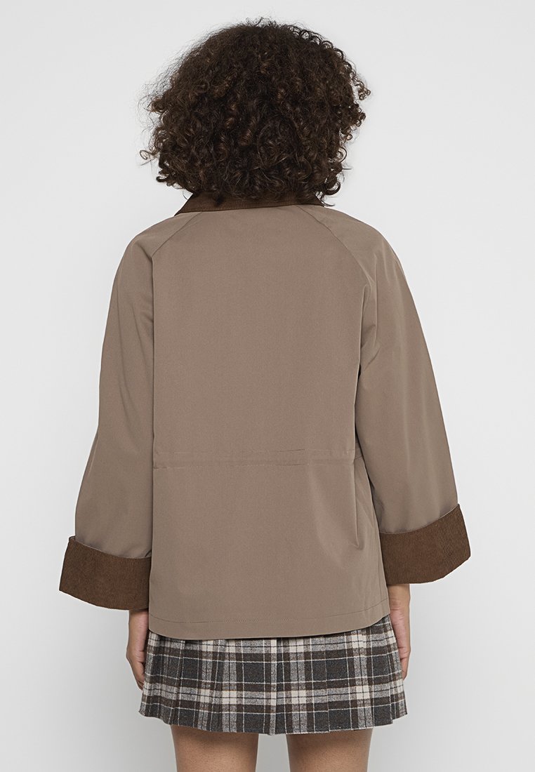 Bruine jas met brede mouwen en een corduroy accent bij de manchetten, gemaakt van een gladde stof. Gedragen over een geruite rok.