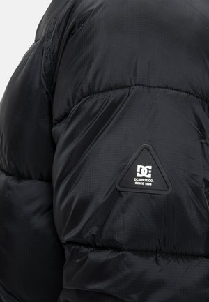 Čierna páperová bunda s prešívanou textúrou, s trojuholníkovou nášivkou na pleci, vyrobená z lesklého, ľahkého materiálu.