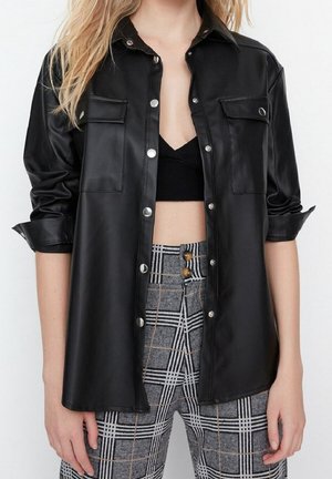 Vrouw draagt een zwarte nepleren blouse met knopen over een zwart crop top en grijze geruite high-waist broek met bruine knopen.