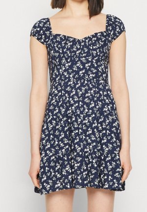 Robe courte bleu marine à fleurs avec manches courtes, corsage ajusté, décolleté en cœur et jupe plissée portée par une femme.