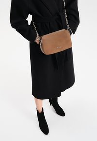 Brązowa skórzana torebka na łańcuchu, prostokątny kształt, zamek błyskawiczny i metalowe logo. Noszona z czarnym płaszczem i czarnymi botkami za kostkę.