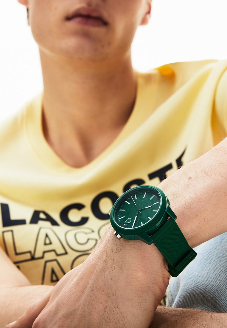 Persona vistiendo una camiseta amarilla de Lacoste y un reloj de pulsera verde con una esfera redonda simple y correa de silicona.