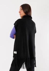 Poncho nero frangiato in tessuto morbido con colletto alto, dotato di etichetta. Design senza maniche che presenta un aspetto a strati con un colore contrastante sottostante.