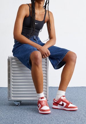 Persoon in een zwart hemdje, denim knielange shorts, witte sokken en rood-witte Nike sneakers zittend op een zilveren koffer met wieltjes.