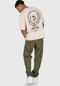 Beige, túlságosan nagy t-shirt, grafikával a hátsó részén; olívazöld cargo nadrág, számos zsebbel; fehér sportcipő. Egyszerű, laza öltözet.