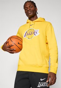 Sweatshirt amarela com o logótipo dos "Los Angeles Lakers" em roxo e branco, com bolso canguru, combinada com calças de moletom pretas apresentando um logótipo semelhante.