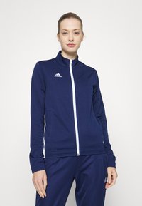 Dunkelblaue Zip-Jacke aus weichem Stoff mit hohem Kragen, ausgestattet mit einem weißen Reißverschluss und kleinem weißen Logo auf der linken Brust.