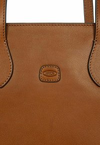 Sac fourre-tout en cuir marron avec une texture lisse, des bords cousus et un patch ovale de marque. Dispose de longues poignées et d'un design minimaliste.