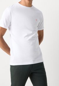 Mann trägt ein einfarbiges weißes T-Shirt mit kurzen Ärmeln und einem kleinen roten "II"-Logo auf der Brust sowie eine dunkelgrüne Hose. Er steht vor einem weißen Hintergrund.