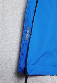 Gros plan sur une fermeture éclair de veste bleue sur un tissu gris avec une petite étiquette réfléchissante portant la mention "RAIN.RDY" près de la couture de la fermeture éclair.